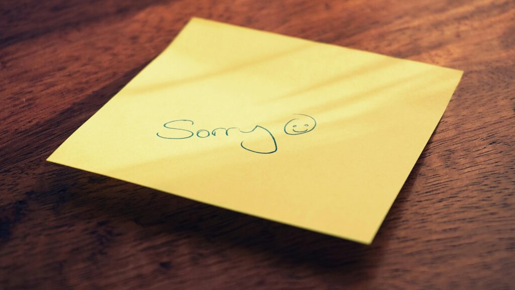 Gelber Haftnotizzettel auf einer Holzoberfläche. Darauf steht handgeschrieben „Sorry“ in grüner Tinte, gefolgt von einem lächelnden Gesicht.