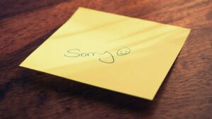 Gelber Haftnotizzettel auf einer Holzoberfläche. Darauf steht handgeschrieben „Sorry“ in grüner Tinte, gefolgt von einem lächelnden Gesicht.