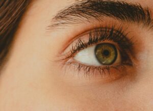 Nahaufnahme eines menschlichen Auges mit grünlich-brauner Iris, sichtbaren Wimpern, Augenbraue und detaillierter Hautstruktur.