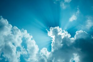 Blauer Himmel mit vereinzelten weißen Wolken, durch die Sonnenstrahlen hindurchscheinen und ein strahlendes Lichtspiel erzeugen.