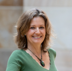 Person mit schulterlangem Haar, grünem Oberteil und auffälliger Halskette mit großem Anhänger. Die Aufnahme wurde im Freien vor einem neutralen, unscharfen Hintergrund gemacht.