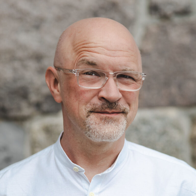 Männliche Person, trägt eine Brille und ein weißes Hemd mit Knöpfen. Im Hintergrund sind steinerne oder betonartige Strukturen in einer Außenumgebung zu sehen.