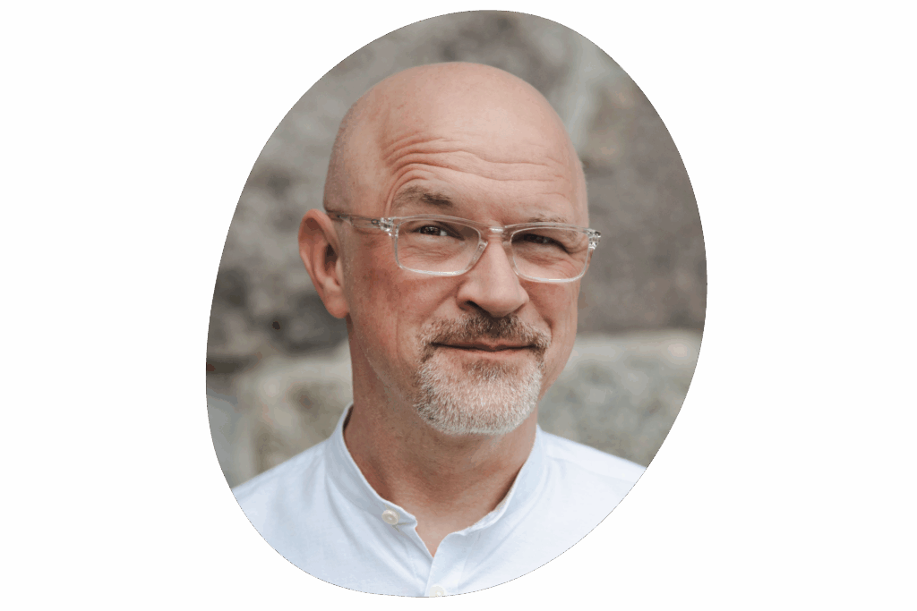 Männliche Person, trägt eine Brille und ein weißes Hemd mit Knöpfen. Im Hintergrund sind steinerne oder betonartige Strukturen in einer Außenumgebung zu sehen.