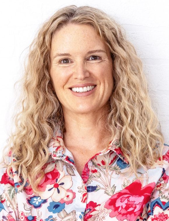 Frau mit langen welligen blonden Haaren. Sie trägt eine Bluse mit pinkem, blauem und beige-farbenem Blumenprint. Sie steht vor einer weißen Wand.