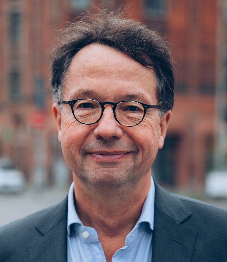 Person mit Brille, trägt ein blaues Hemd und eine dunkle Jacke. Im Hintergrund sind Gebäude und möglicherweise etwas Grün zu sehen – eine städtische Umgebung.
