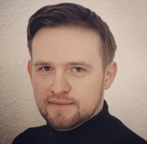 Person mit kurzen, dunklen Haaren vor einem hellen, einfarbigen Hintergrund.