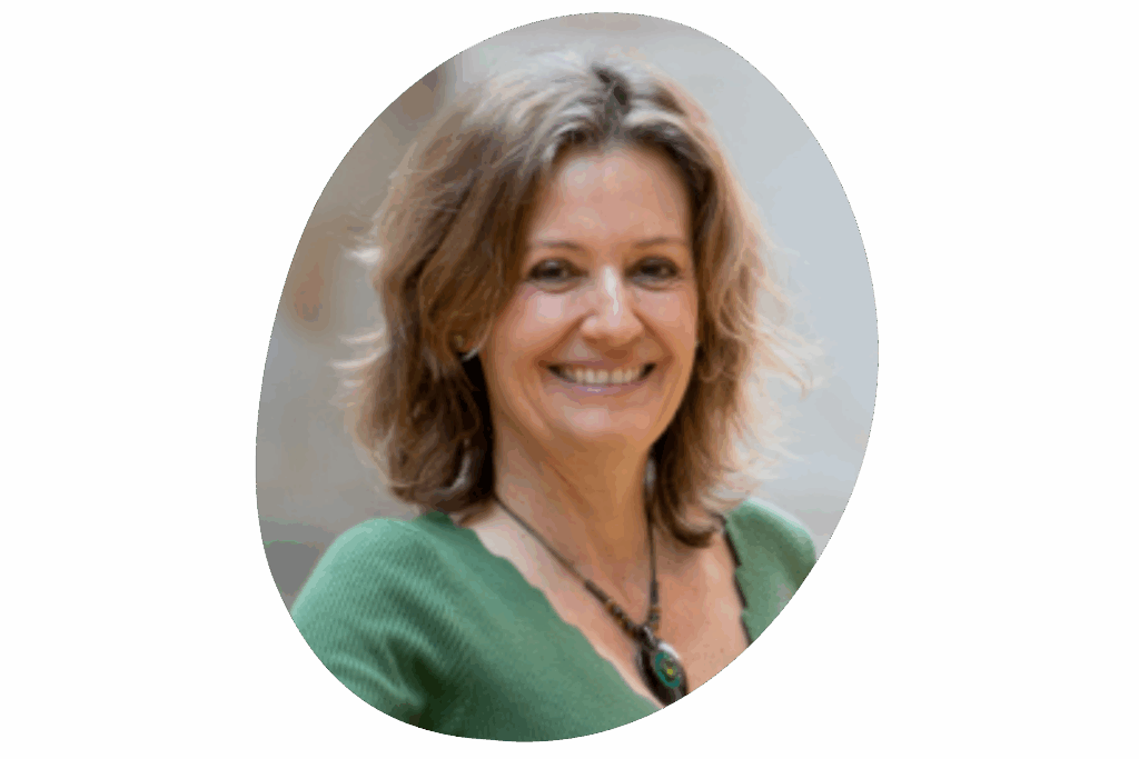 Person mit schulterlangem Haar, grünem Oberteil und auffälliger Halskette mit großem Anhänger. Die Aufnahme wurde im Freien vor einem neutralen, unscharfen Hintergrund gemacht.