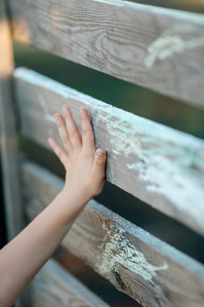Nahaufnahme einer Kinderhand, die eine hölzerne Zaunlatte berührt. Der Zaun besteht aus horizontalen Planken mit sichtbarer Maserung und stellenweise abblätternder Farbe. Der Hintergrund ist unscharf.