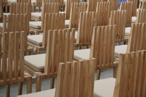 Reihenweise angeordnete Holzstühle mit hellen Sitzpolstern in einem leeren, lichtdurchfluteten Innenraum – vermutlich ein Saal oder Auditorium.