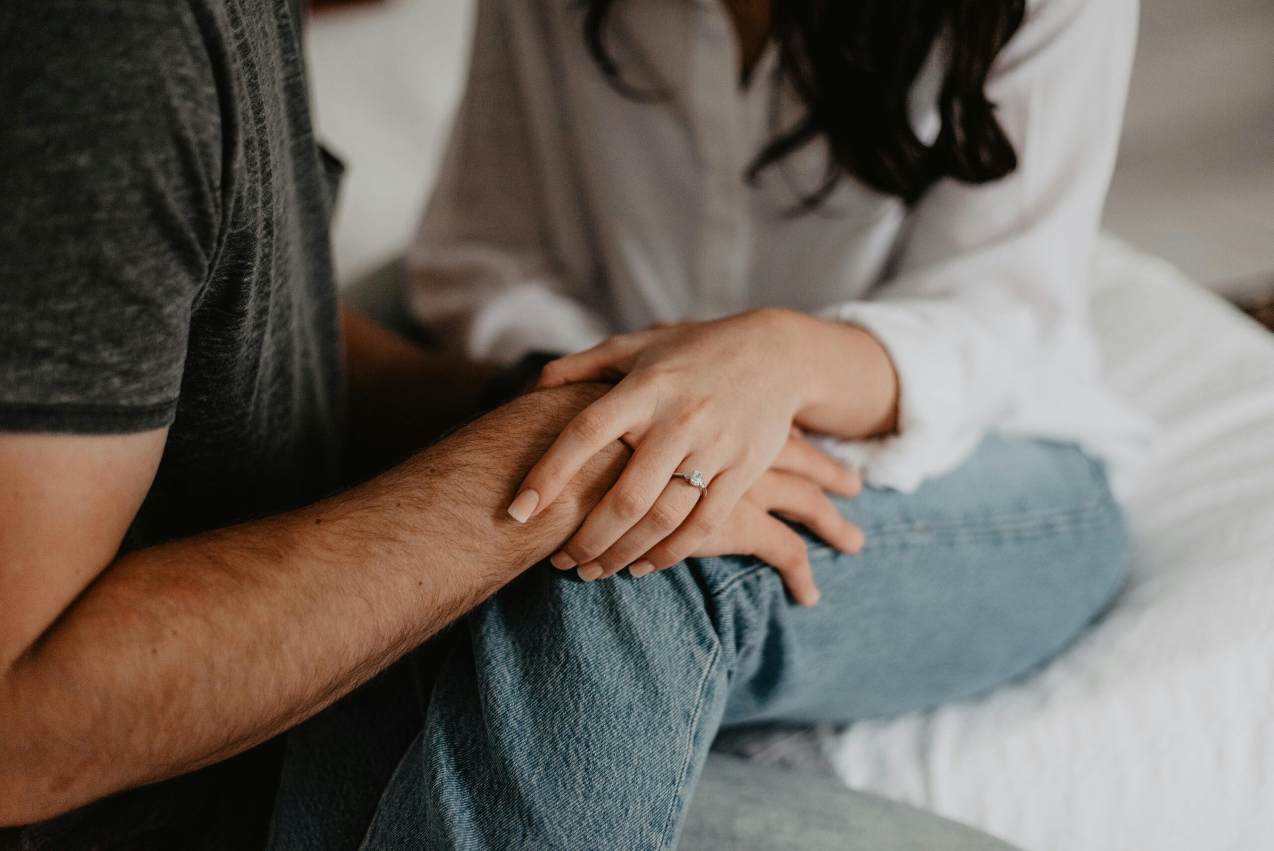 Zwei Personen sitzen eng nebeneinander. Eine Person trägt ein weißes Shirt und blaue Jeans, die andere ein dunkelgraues Shirt. Die Person im weißen Shirt hält mit beiden Händen den Arm der anderen Person. Ein Ring ist sichtbar