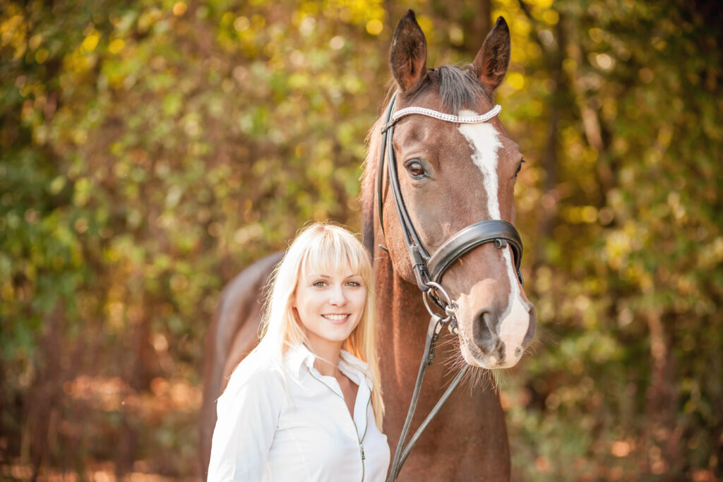 Pferd mit Trense steht neben einer Person in weißem Oberteil vor einem Hintergrund aus herbstlich wirkenden Bäumen.