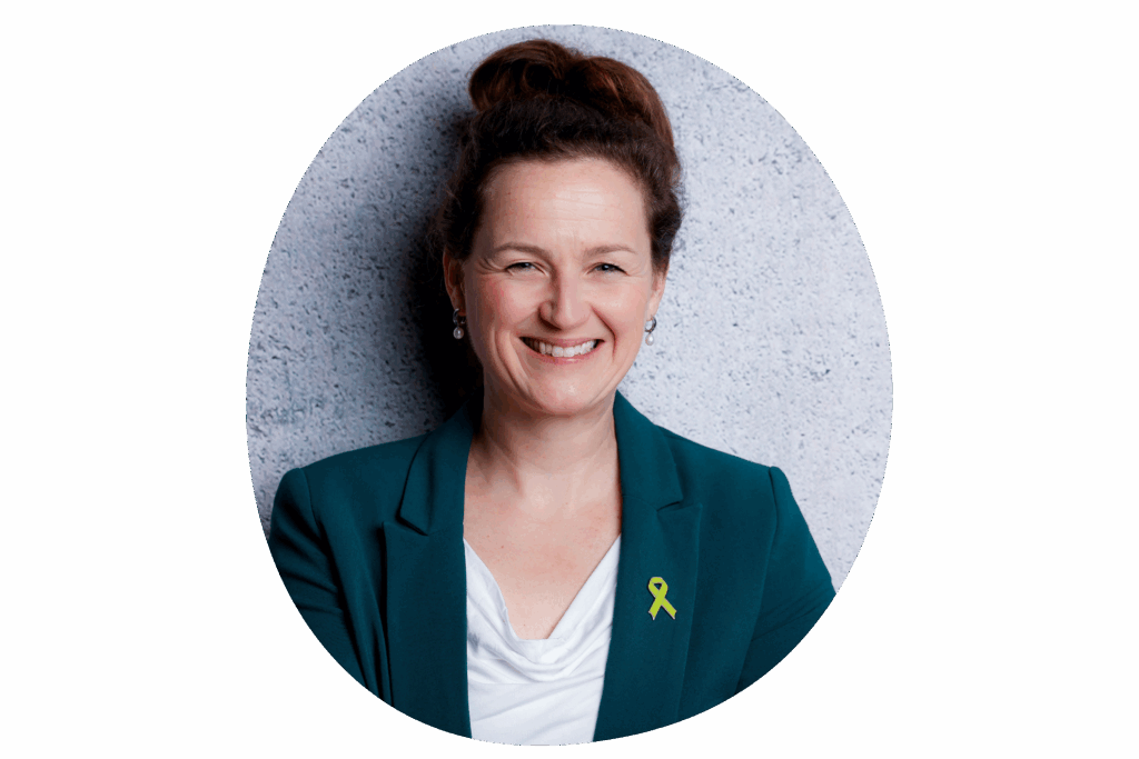 Person in grünem Blazer mit weißem Oberteil, trägt eine Anstecknadel in Form einer grünen Schleife, vor grauer Wand