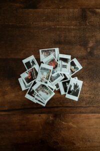 Ein Stapel kleiner Polaroid-Fotos liegt auf einem Holztisch. Die Bilder zeigen verschiedene Motive wie Häuser, Pflanzen und Landschaften.