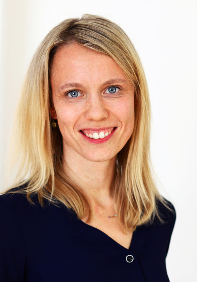 Person mit schulterlangem, blondem Haar trägt eine dunkelblaue Bluse mit kleinem Knopf und steht vor einem hellen, neutralen Hintergrund.