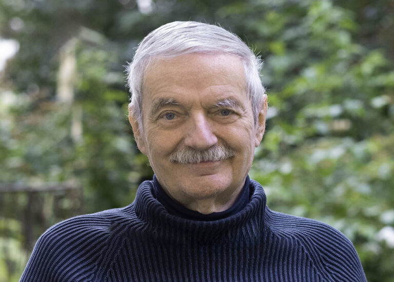 Person mit grauem Haar trägt einen dunkelblauen, gerippten Rollkragenpullover und sitzt im Freien vor einem unscharfen, grünen Hintergrund mit Bäumen und Pflanzen.