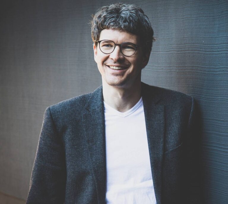 Person mit lockigem, dunkelblondem Haar trägt ein weißes T-Shirt unter einem dunklen Sakko und lehnt an einer strukturierten Wand in einem gedämpften Licht.