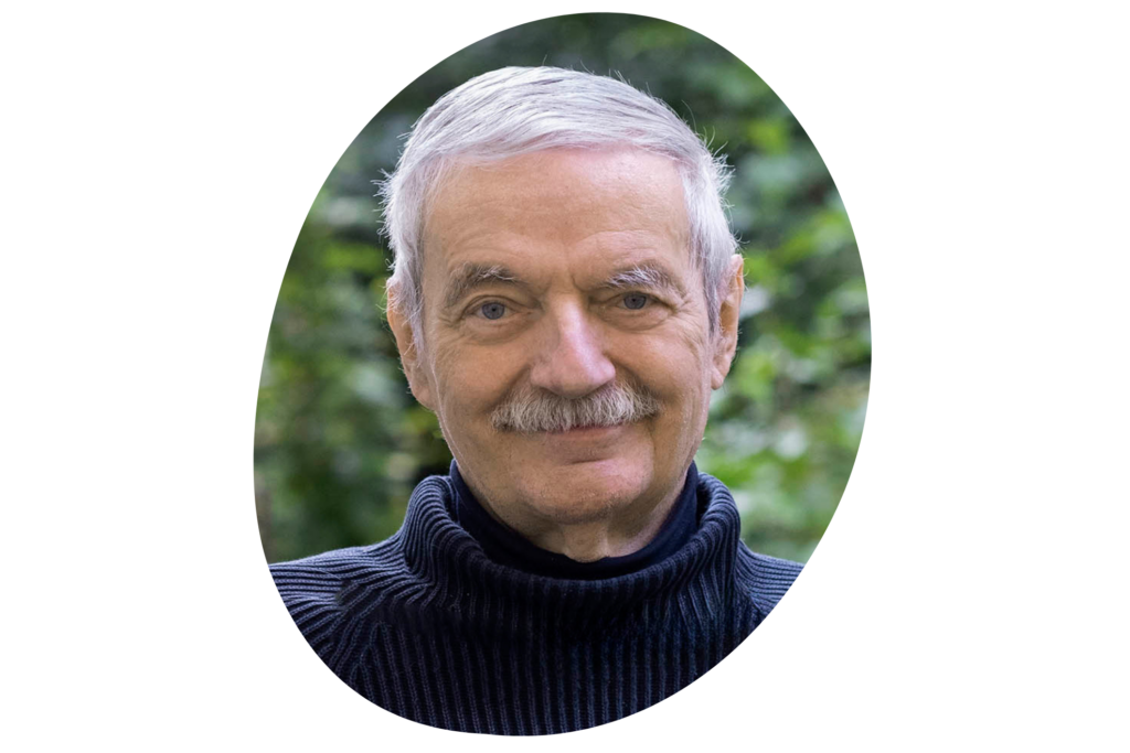 Person mit grauem Haar trägt einen schwarzen Rollkragenpullover. Der Hintergrund zeigt grüne, unscharfe Vegetation.