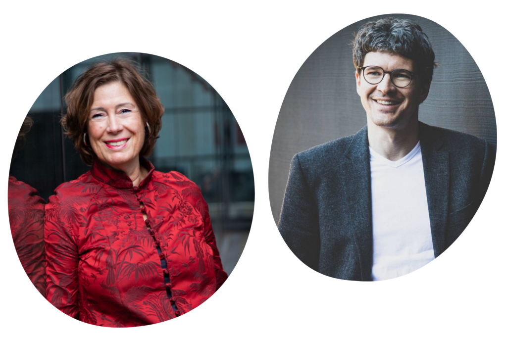 2 Personen: Die Person rechts trägt ein dunkelgraues Sakko über einem weißen T-Shirt. Der Hintergrund ist eine strukturierte, graue Wand. Die Person links trägt eine rote, gemusterte Bluse mit asiatisch inspiriertem Stehkragen und Knopfleiste. Im Hintergrund ist eine spiegelnde Glasfläche zu sehen.