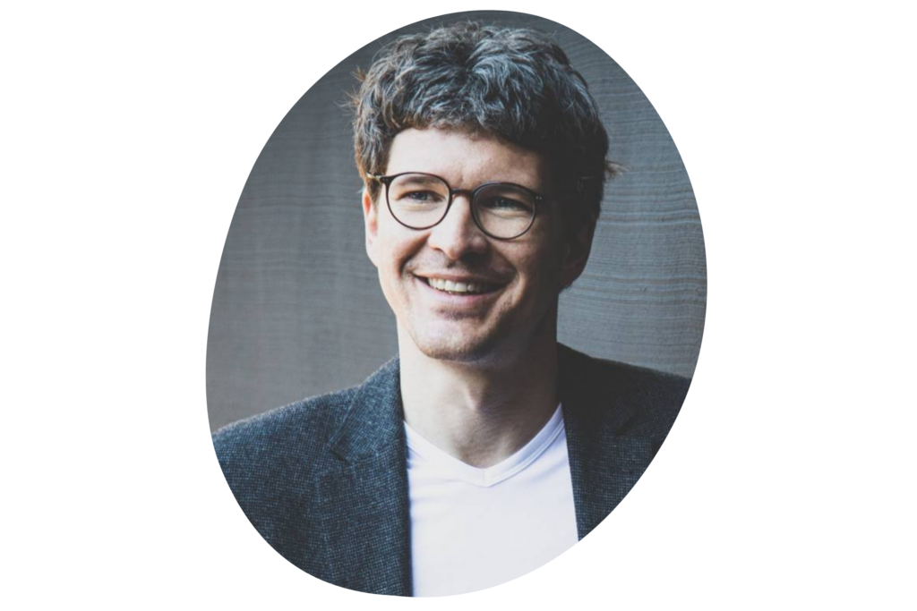 Person trägt ein dunkelgraues Sakko über einem weißen T-Shirt. Der Hintergrund ist eine strukturierte, graue Wand.