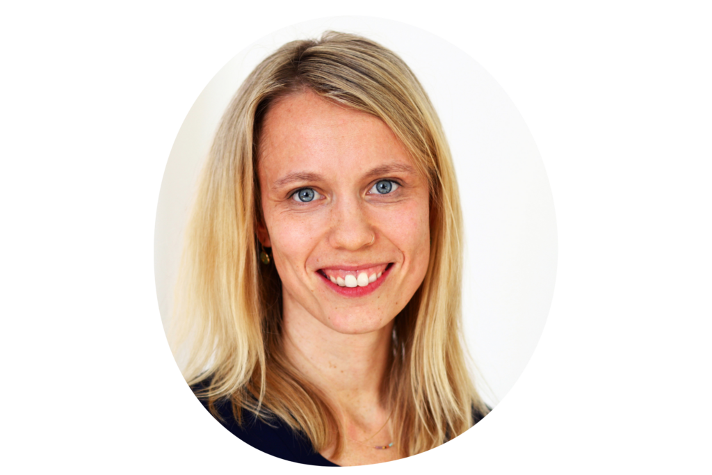 Person mit schulterlangem, blondem Haar trägt ein dunkelblaues Oberteil. Der Hintergrund ist ein neutraler, heller Farbton.