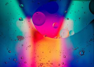 Foto einer mit Tropfen bedeckten Glasscheibe vor einem unscharfen, verschiedenfarbig beleuchtenden Hintergrund.