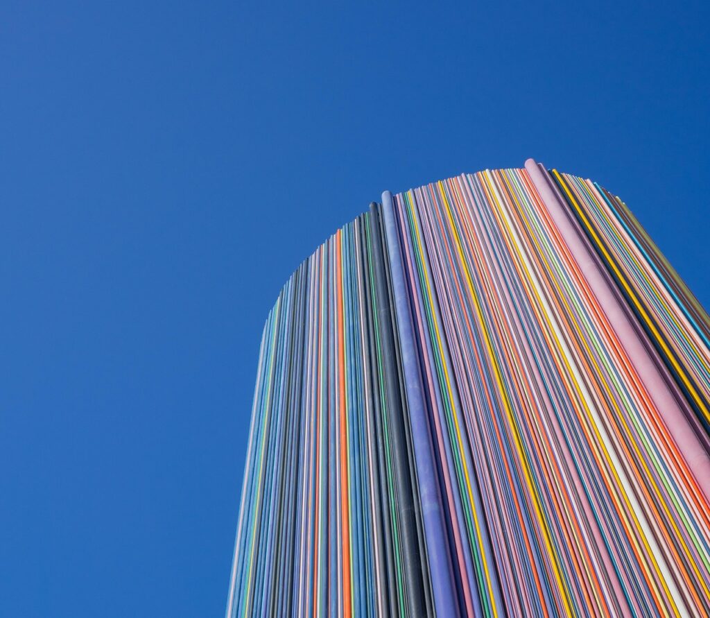 Hoher, runder Gebäudeturm, der vollständig aus vertikal verlaufenden, farbigen Streifen besteht, vor klarem blauem Himmel. Die bunten Linien wirken wie dicht nebeneinander angeordnete Farbtafeln und erzeugen einen dynamischen, modernen Eindruck.