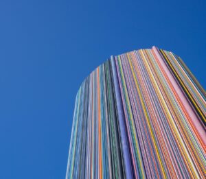 Hoher, runder Gebäudeturm, der vollständig aus vertikal verlaufenden, farbigen Streifen besteht, vor klarem blauem Himmel. Die bunten Linien wirken wie dicht nebeneinander angeordnete Farbtafeln und erzeugen einen dynamischen, modernen Eindruck.
