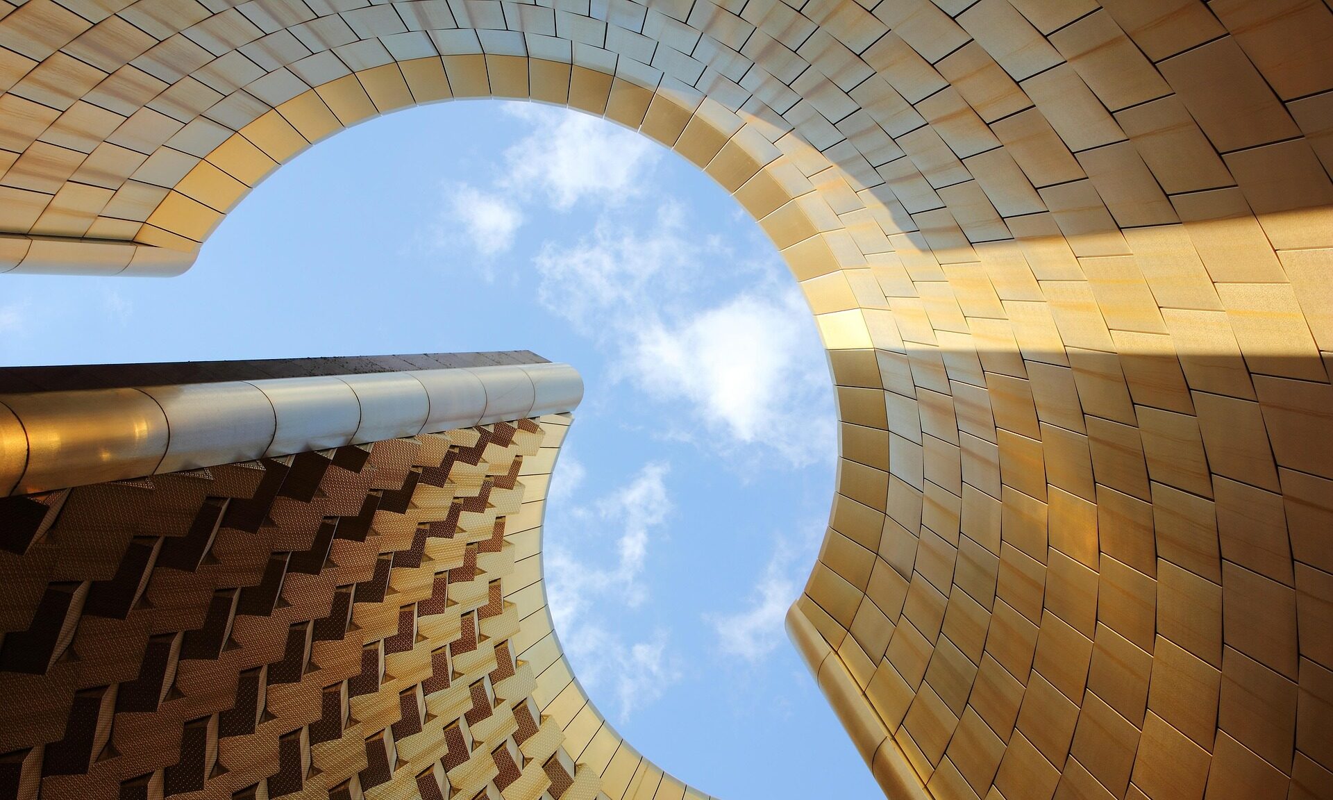 Blick von unten durch eine große, runde Öffnung in einer goldfarben verkleideten Gebäudeform hinauf in den blauen Himmel mit einigen Wolken.