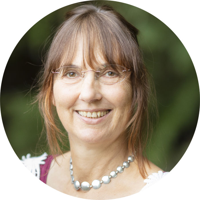 Nahaufnahme des Gesichts einer Frau, die braune Haare hat, eine Brille und auffällige Perlenkette trägt.