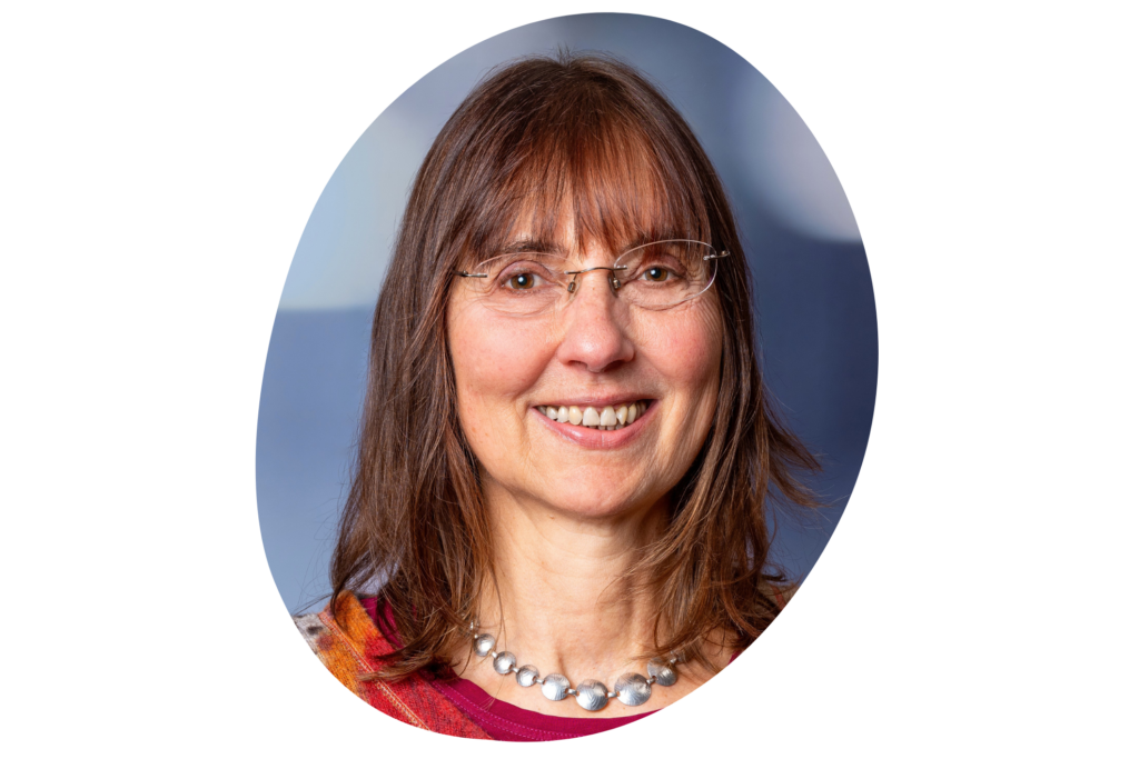 Nahaufnahme des Gesichts einer Frau vor einem dunklen Hintergrund, die braune Haare hat, eine Brille und auffällige Perlenkette trägt.