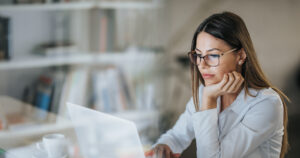 Person sitzt an einem Schreibtisch vor einem Laptop, stützt den Kopf auf eine Hand und arbeitet in einer modernen, hellen Büro- oder Arbeitsumgebung; im Vordergrund ist ein weißes Getränkeglas zu sehen.