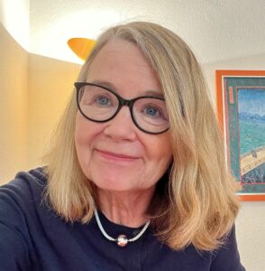 Eine Frau mit schulterlangen blonden Haaren, die eine Brille mit einem dickeren schwarzen Rand trägt und lächelt. Im Hintergrund ist eine Lampe und ein eingerahmtes Bild zu sehen.