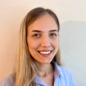 Porträt einer Person mit langen, hellen Haaren, frontal fotografiert vor hellem, unscharfem Hintergrund, in einem hellblauen Hemd.