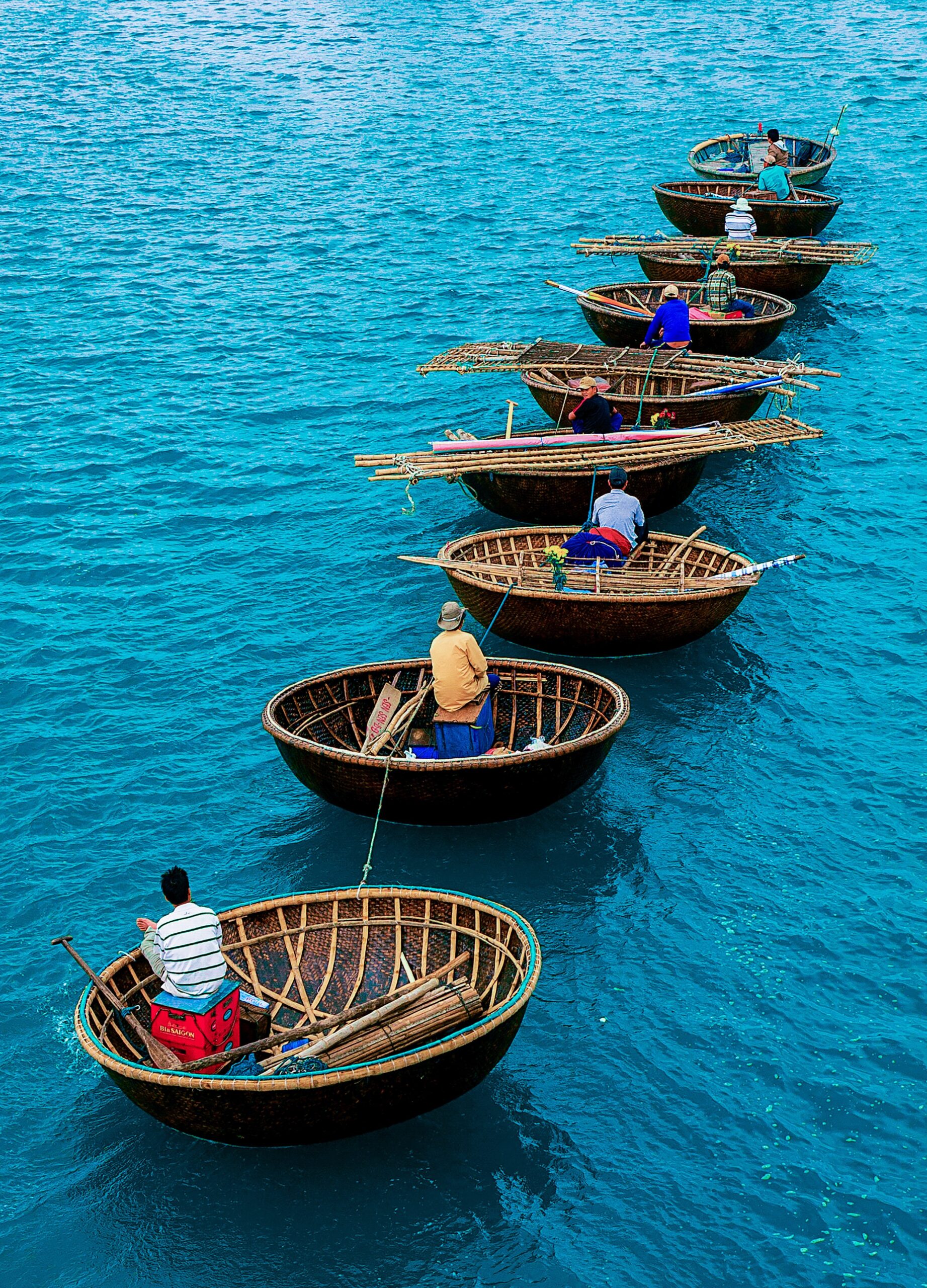 Mehrere runde, geflochtene Korbboote mit sitzenden Personen auf türkisblauem Wasser hintereinander angeordnet.