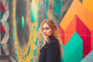 Junge, langhaarige blonde Frau vor bunter Wand im Freien.