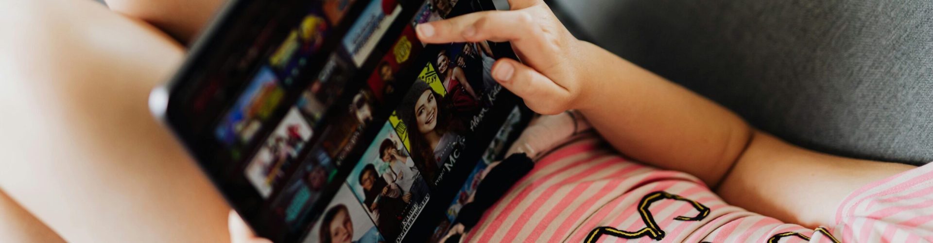 Ein Mädchen liegt auf einem grauen Sofa und hält ein Tablet in der Hand. Auf dem Bildschirm sind Vorschaubilder von Filmen und Serien zu erkennen, was auf die Nutzung eines Streaming-Dienstes schließen lässt. Sie trägt ein rosa-weiß gestreiftes Shirt mit teilweise lesbarem Aufdruck.