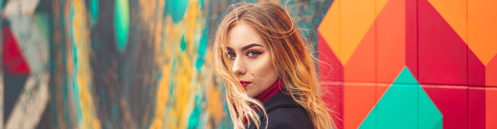 Junge, langhaarige blonde Frau vor bunter Wand im Freien.