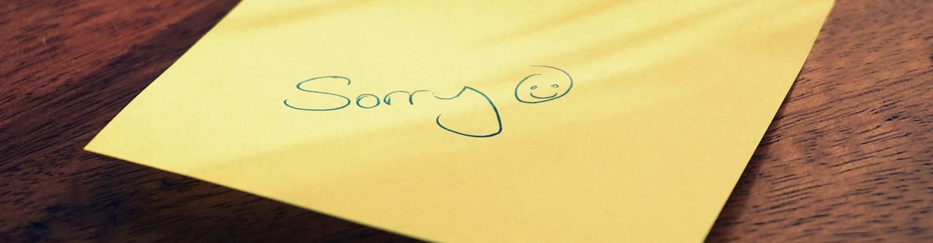 Gelber Haftnotizzettel auf einer Holzoberfläche. Darauf steht handgeschrieben „Sorry“ in grüner Tinte, gefolgt von einem lächelnden Gesicht.