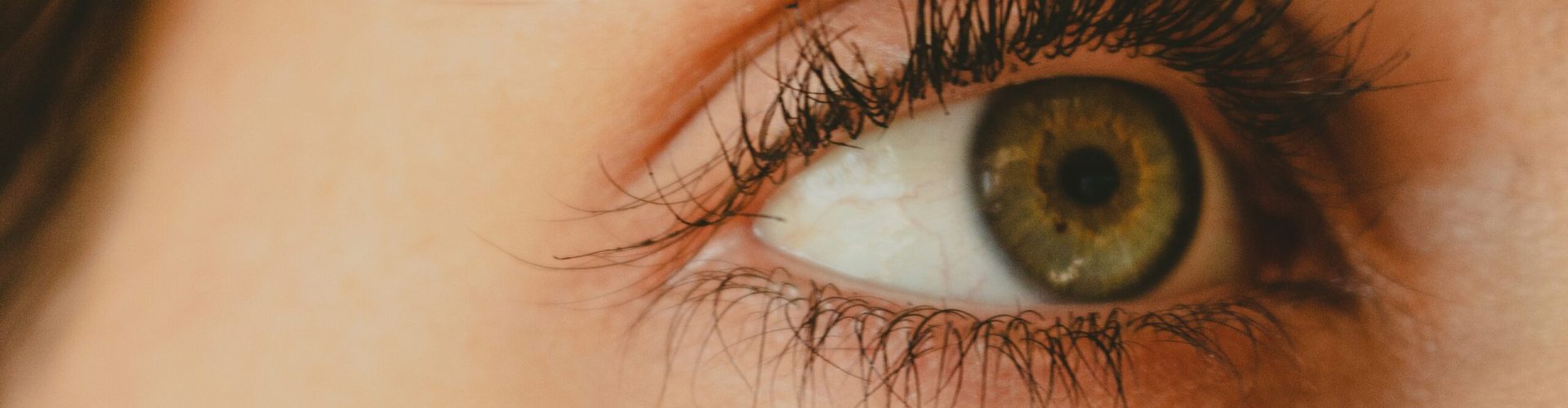 Nahaufnahme eines menschlichen Auges mit grünlich-brauner Iris, sichtbaren Wimpern, Augenbraue und detaillierter Hautstruktur.
