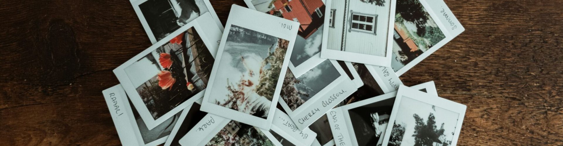 Ein Stapel kleiner Polaroid-Fotos liegt auf einem Holztisch. Die Bilder zeigen verschiedene Motive wie Häuser, Pflanzen und Landschaften.