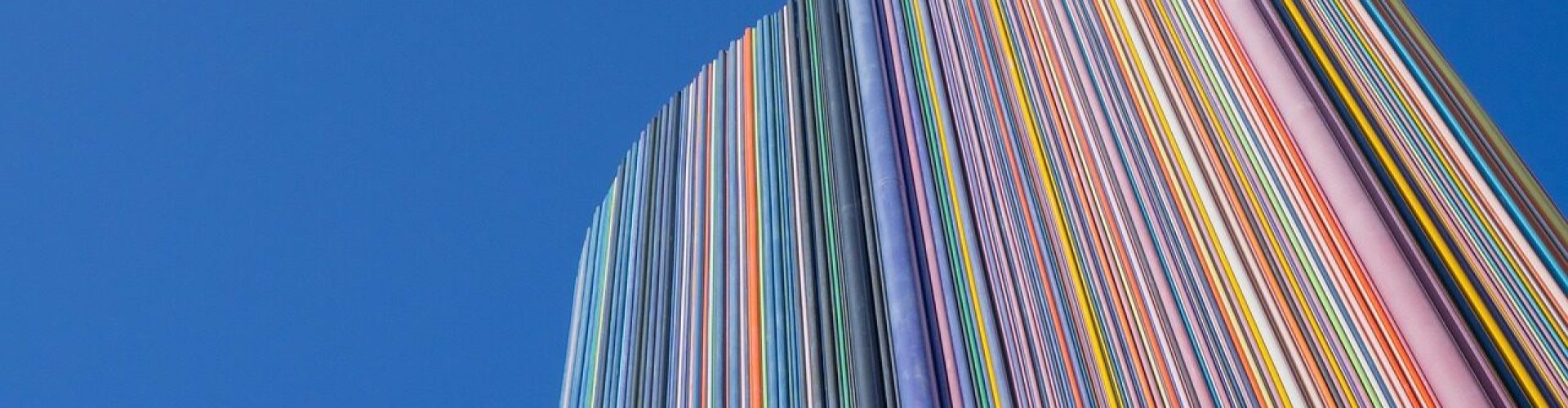 Hoher, runder Gebäudeturm, der vollständig aus vertikal verlaufenden, farbigen Streifen besteht, vor klarem blauem Himmel. Die bunten Linien wirken wie dicht nebeneinander angeordnete Farbtafeln und erzeugen einen dynamischen, modernen Eindruck.