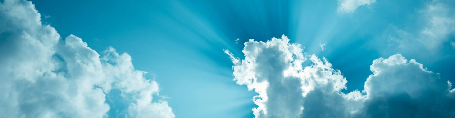 Blauer Himmel mit vereinzelten weißen Wolken, durch die Sonnenstrahlen hindurchscheinen und ein strahlendes Lichtspiel erzeugen.