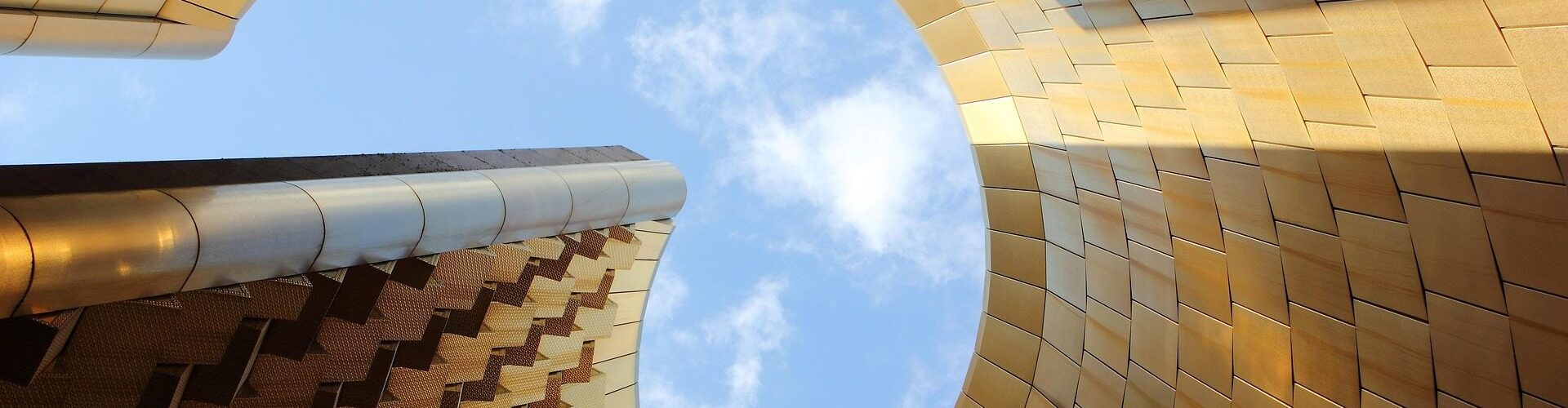 Blick von unten durch eine große, runde Öffnung in einer goldfarben verkleideten Gebäudeform hinauf in den blauen Himmel mit einigen Wolken.