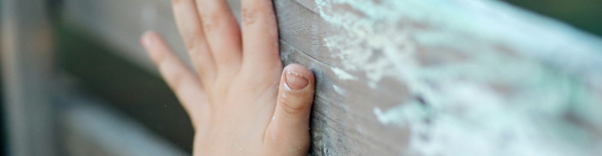 Nahaufnahme einer Kinderhand, die eine hölzerne Zaunlatte berührt. Der Zaun besteht aus horizontalen Planken mit sichtbarer Maserung und stellenweise abblätternder Farbe. Der Hintergrund ist unscharf.