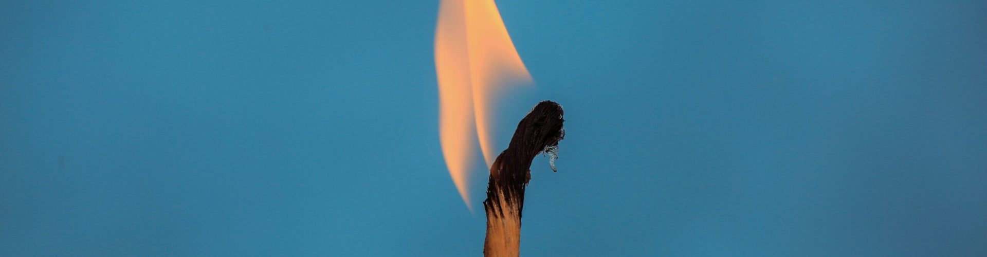 Nahaufnahme eines brennenden Streichholzes vor einem einfarbig blauen Hintergrund. Die Flamme ist gelb-orange und leicht nach links geneigt, während der obere Teil des Streichholzes schwarz verkohlt ist.