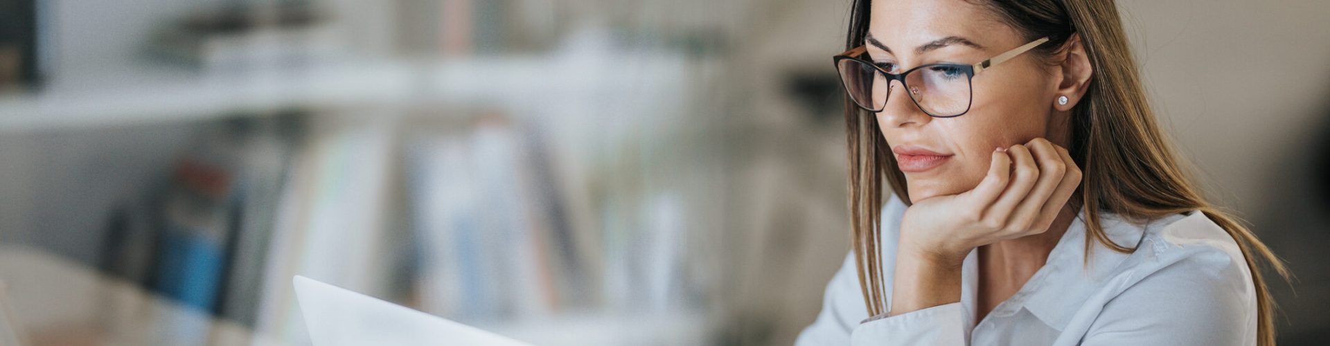 Person sitzt an einem Schreibtisch vor einem Laptop, stützt den Kopf auf eine Hand und arbeitet in einer modernen, hellen Büro- oder Arbeitsumgebung; im Vordergrund ist ein weißes Getränkeglas zu sehen.