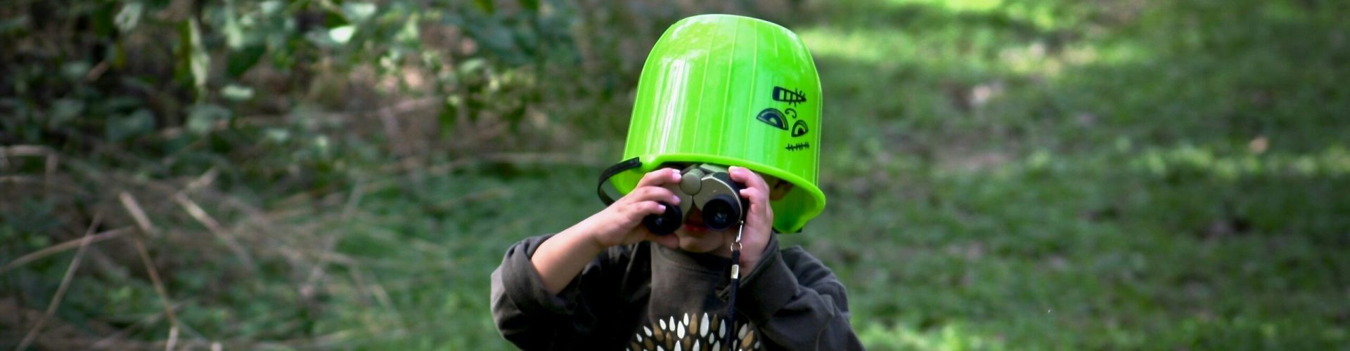 Kind mit Fernglas in der Natur. Es trägt einen grünen Eimer auf dem Kopf.