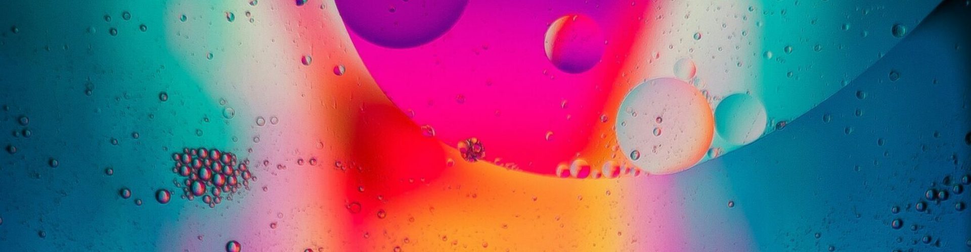 Foto einer mit Tropfen bedeckten Glasscheibe vor einem unscharfen, verschiedenfarbig beleuchtenden Hintergrund.