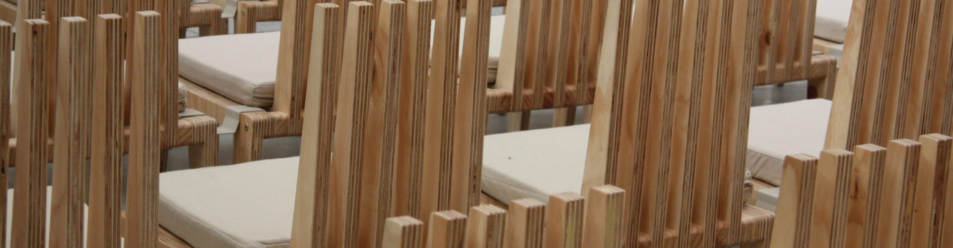 Reihenweise angeordnete Holzstühle mit hellen Sitzpolstern in einem leeren, lichtdurchfluteten Innenraum – vermutlich ein Saal oder Auditorium.