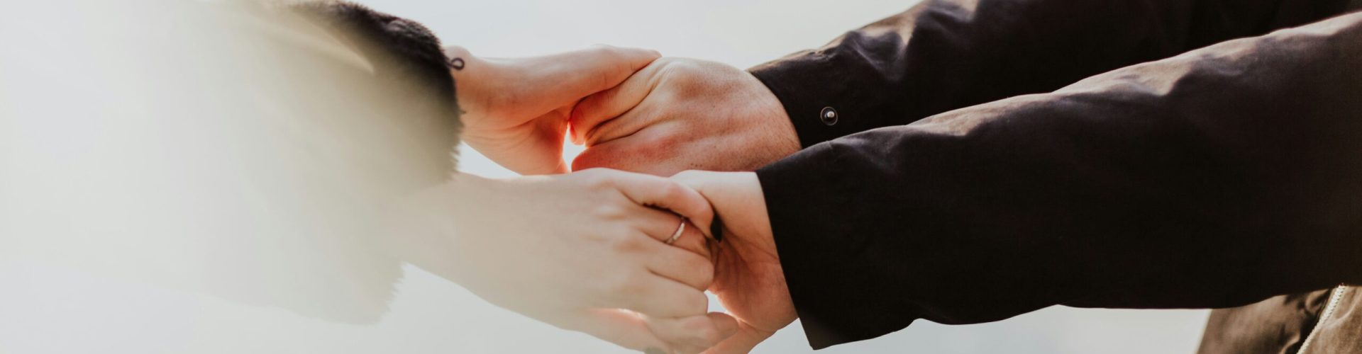 Zwei Personen stehen einander gegenüber und halten sich sanft an den Händen. Beide tragen dunkle Jacken, und der Hintergrund ist hell und unscharf, was eine ruhige, minimalistische Stimmung erzeugt.